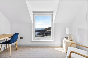 A room with a sloped ceiling, a wooden desk, a blue chair, a bedside table with a lamp, and a view of the sea through a window. - Beautiful 4-bedroom house with stunning sea views in the village of Guileen (Whitegate)