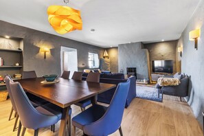 A modern living room with a wooden dining table, blue chairs, a sofa, and a TV. - Beautiful 4-bedroom house with stunning sea views in the village of Guileen (Whitegate)