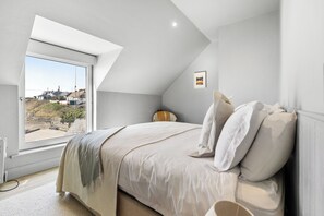 A bedroom with a large window, a bed with white bedding, and a view of a town. - Beautiful 4-bedroom house with stunning sea views in the village of Guileen (Whitegate)