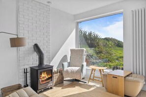 A cozy room with a fireplace, a comfortable chair, a small table, and a view of greenery outside. - Beautiful 4-bedroom house with stunning sea views in the village of Guileen (Whitegate)