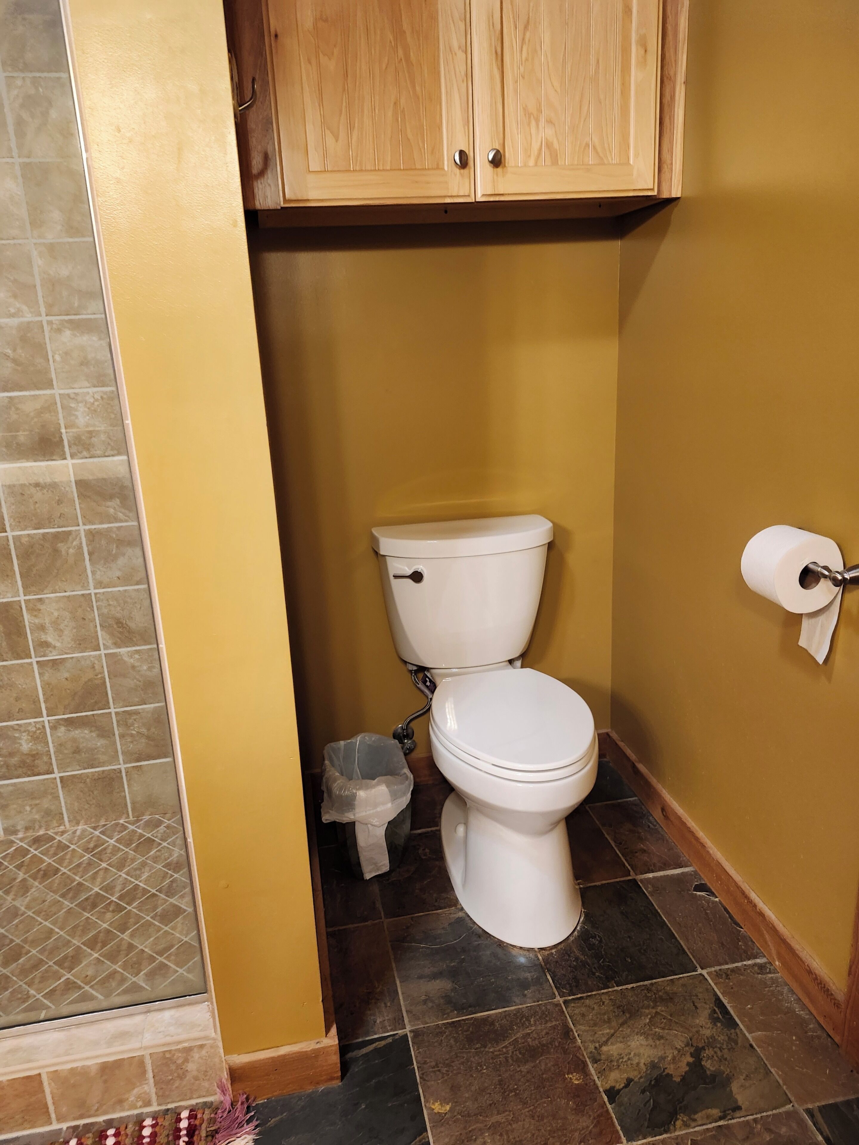 A bathroom with a toilet, a roll of toilet paper, a trash bin, and wooden cabinets.