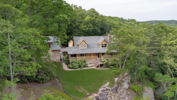 Une grande maison entourée d’une forêt dense, avec une pelouse bien entretenue et un mur de soutènement en pierre.