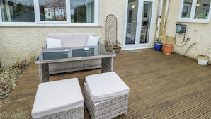 A patio with a wicker sofa, two white cushions, and a small table. There are potted plants and a hose on the patio. - Hafan Hapus (Beaumaris)