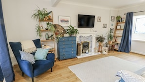 A living room with a blue armchair, wooden floor, a fireplace, and a TV mounted on the wall. - Hafan Hapus (Beaumaris)