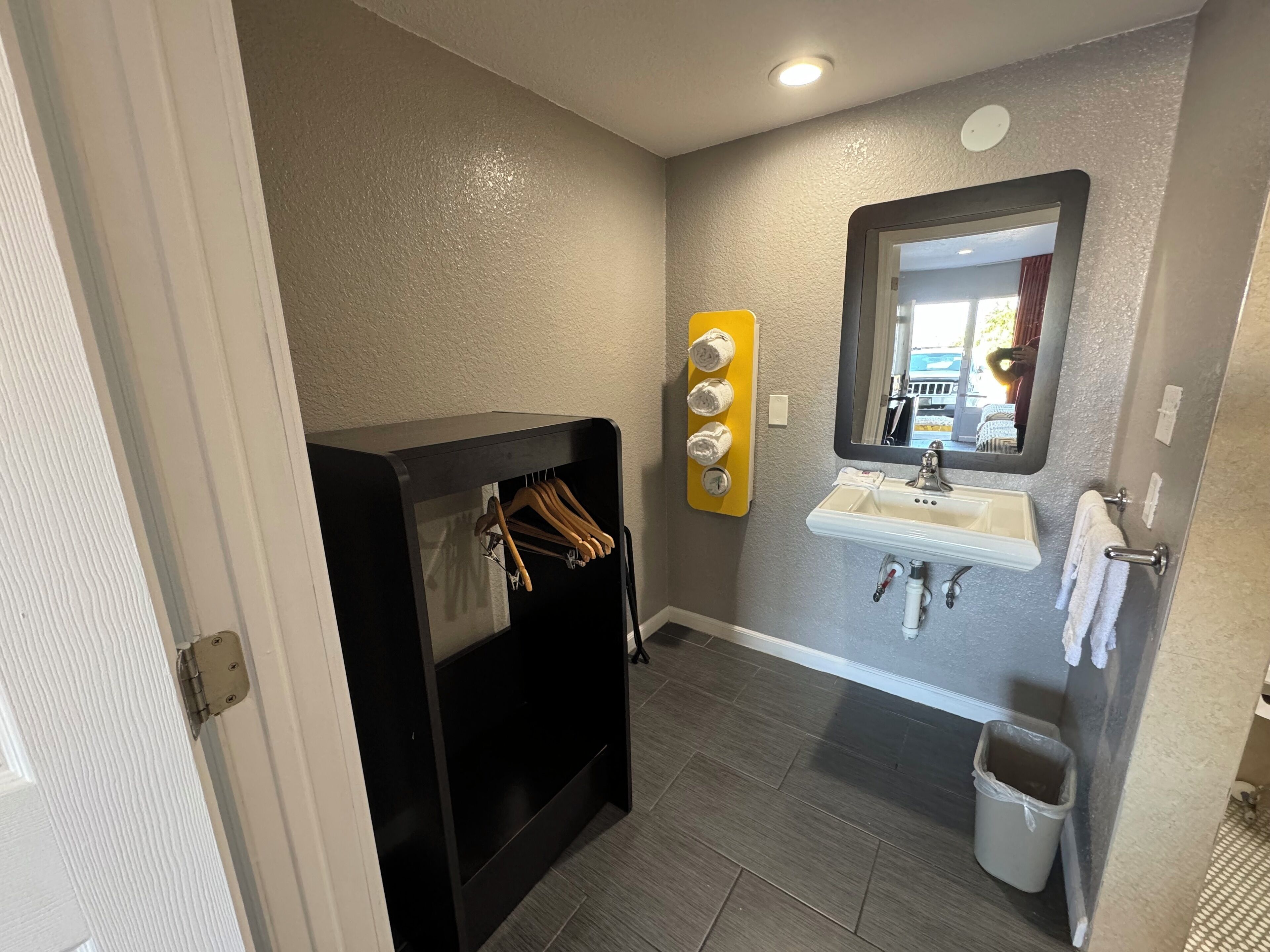 Un bagno con lavandino, specchio, porta asciugamani e un mobile contenitore nero con abiti appesi.