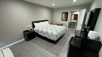 A hotel room with a bed, a desk, a chair, a mirror, a sink, and a trash bin.