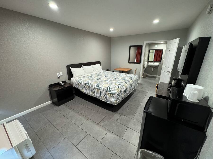 A hotel room with a bed, a desk, a chair, a mirror, a sink, and a trash bin.
