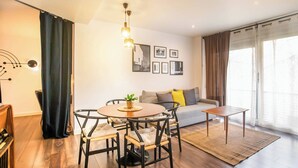 A modern living room with a round dining table, two chairs, a sofa, and a coffee table. There are framed pictures on the wall, a hanging light fixture, and a window with curtains. - Ebe1par1133- Apartment in Avenida Paralelo (Barcelona)