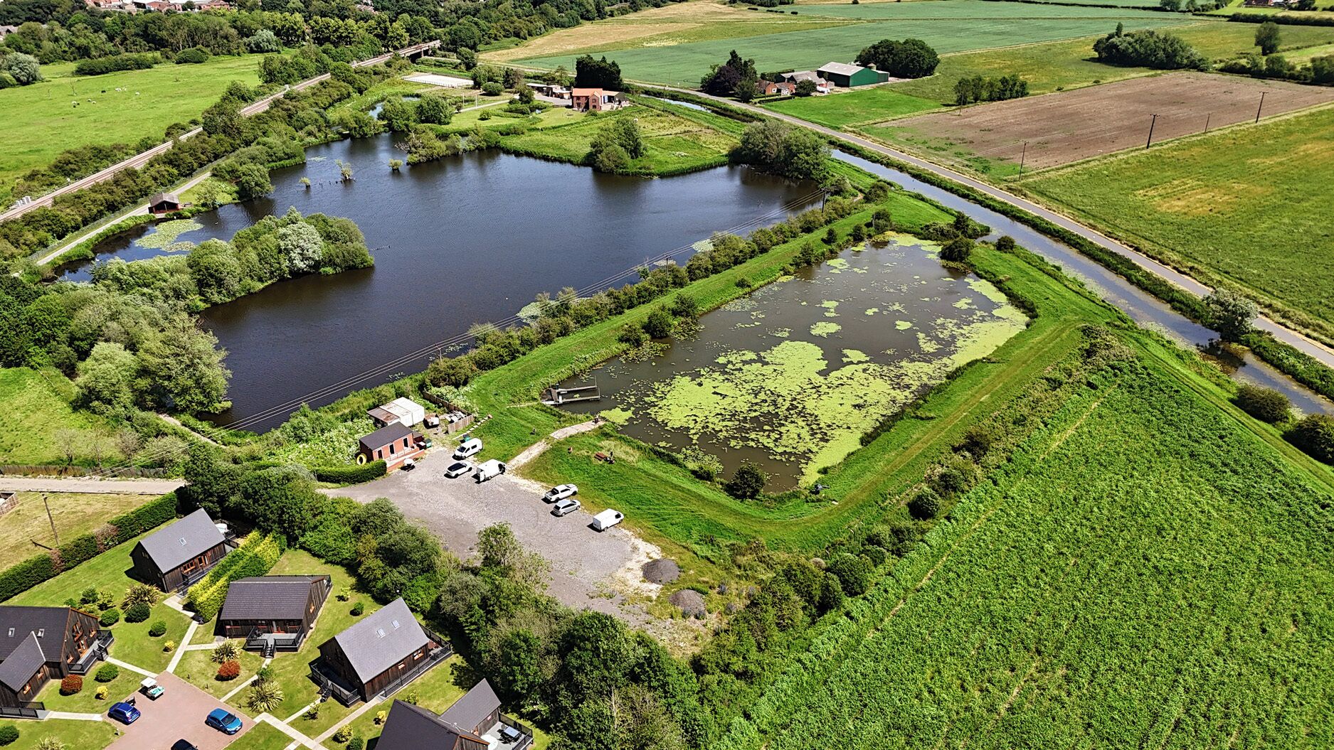 An aerial view of a rural area with a river, green fields, and a small settlement with houses and parked vehicles.