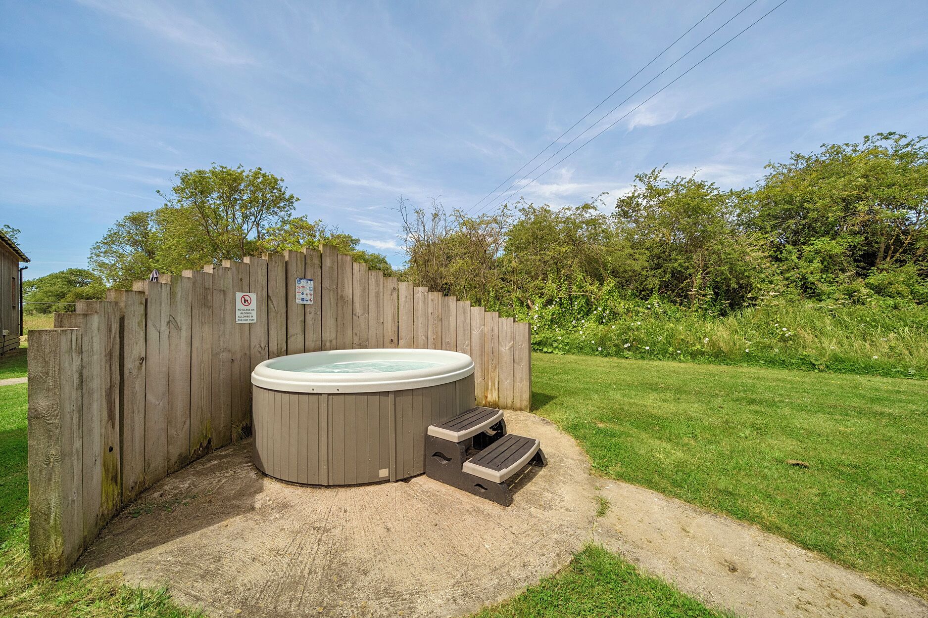 A hot tub with steps, surrounded by a wooden fence, in a grassy yard with trees in the background.