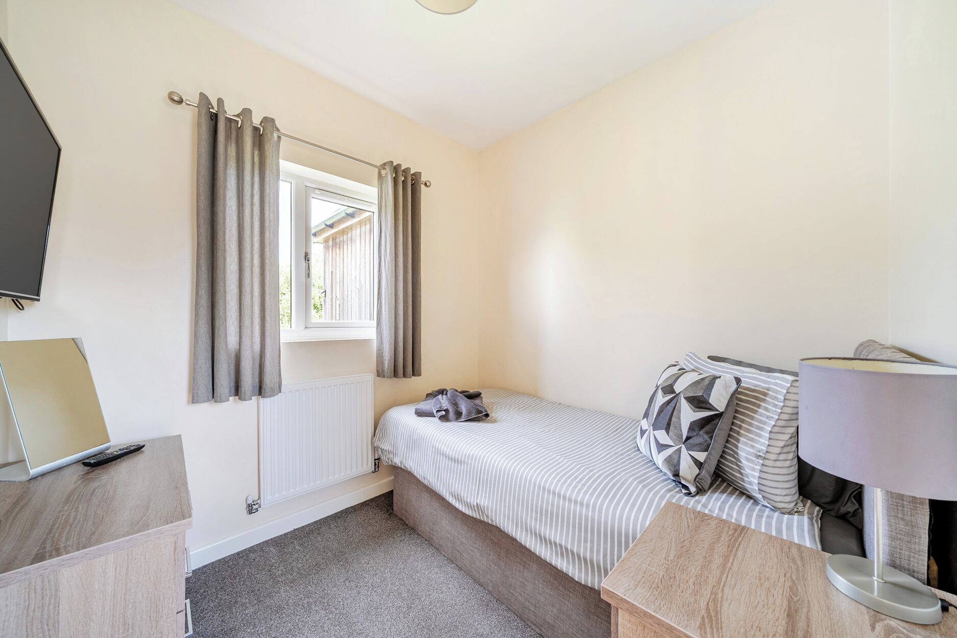 A bedroom with a bed, a window with curtains, a bedside table, a lamp, and a radiator.