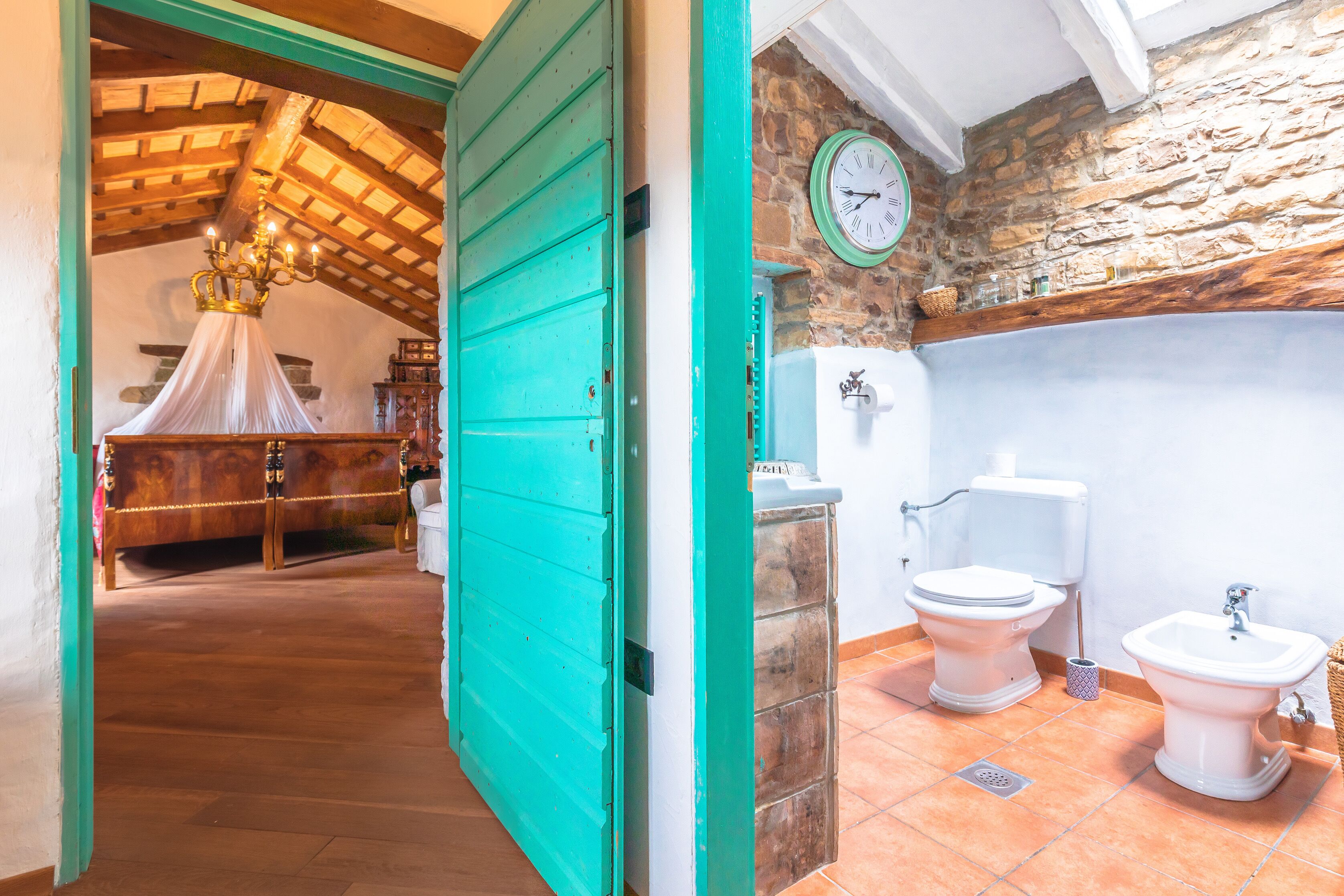 A room with a wooden ceiling, a bed with a canopy, a bathroom with two toilets and a bidet, and a stone wall.