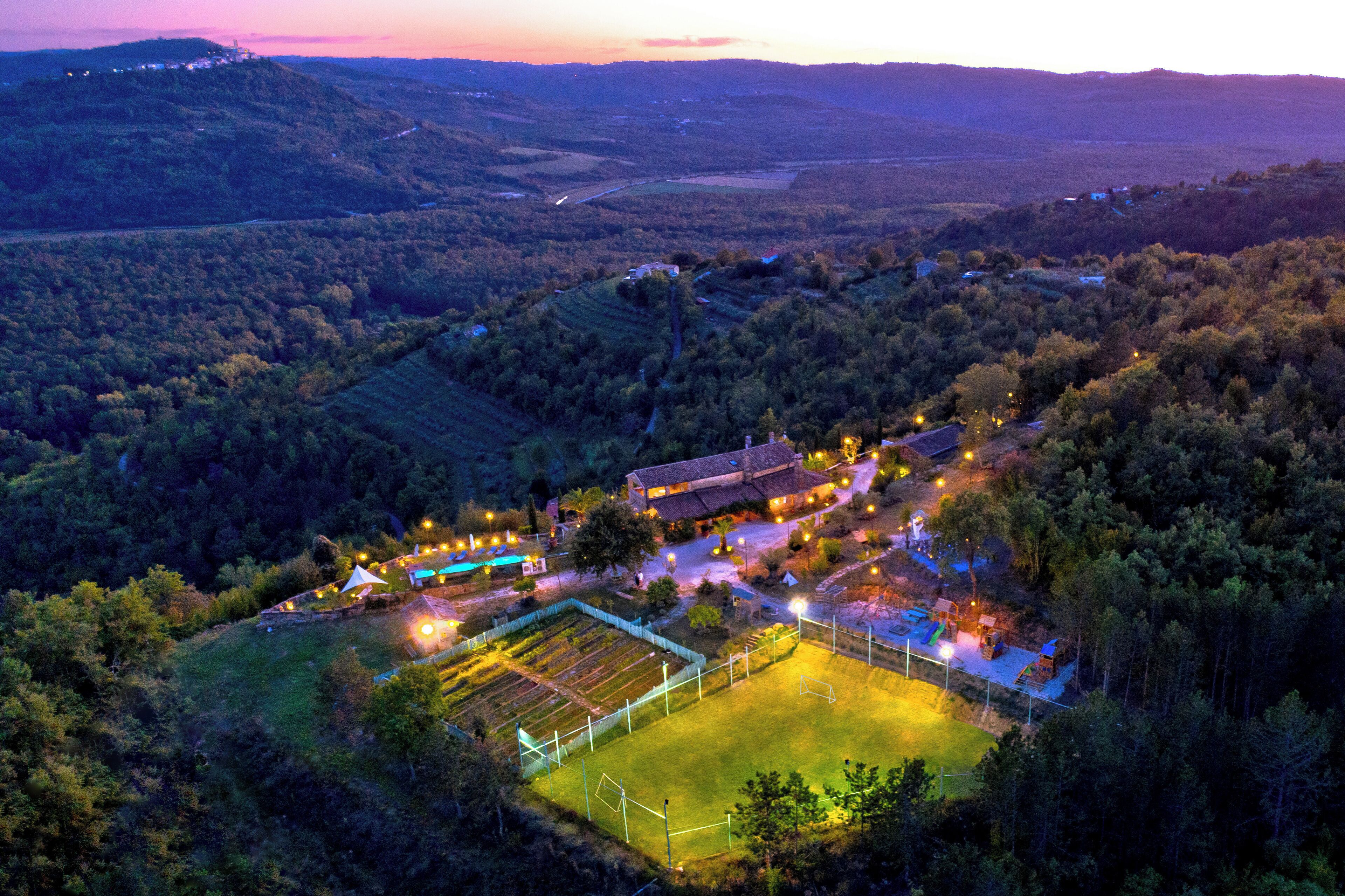 An aerial view of a rural property with a large field, a building with multiple rooms, and surrounding forested hills.