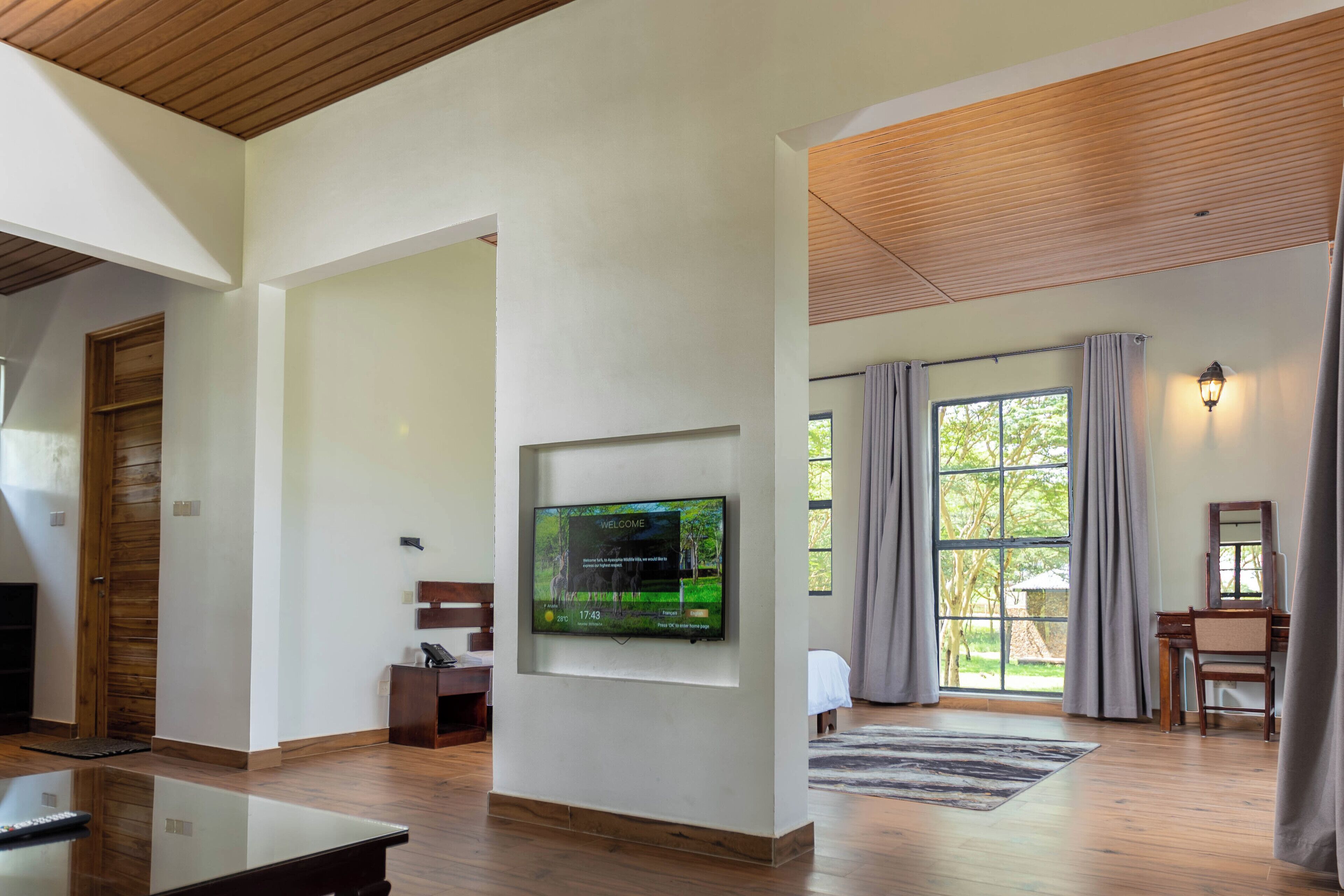 A spacious living room with a large flat-screen TV mounted on the wall, a wooden ceiling, and a large window with curtains.