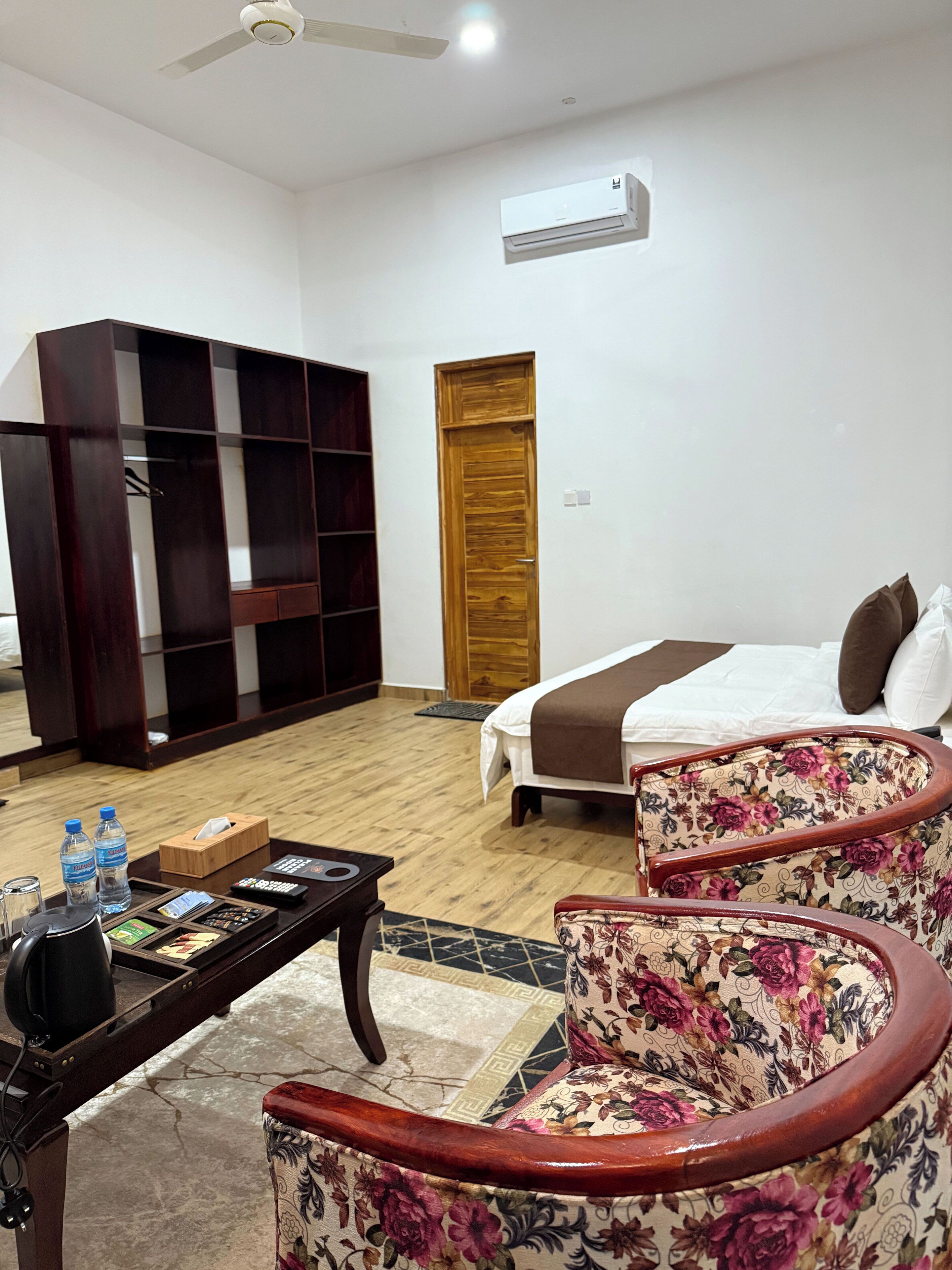 A hotel room with a bed, a wooden wardrobe, a desk with a telephone, and two floral armchairs.