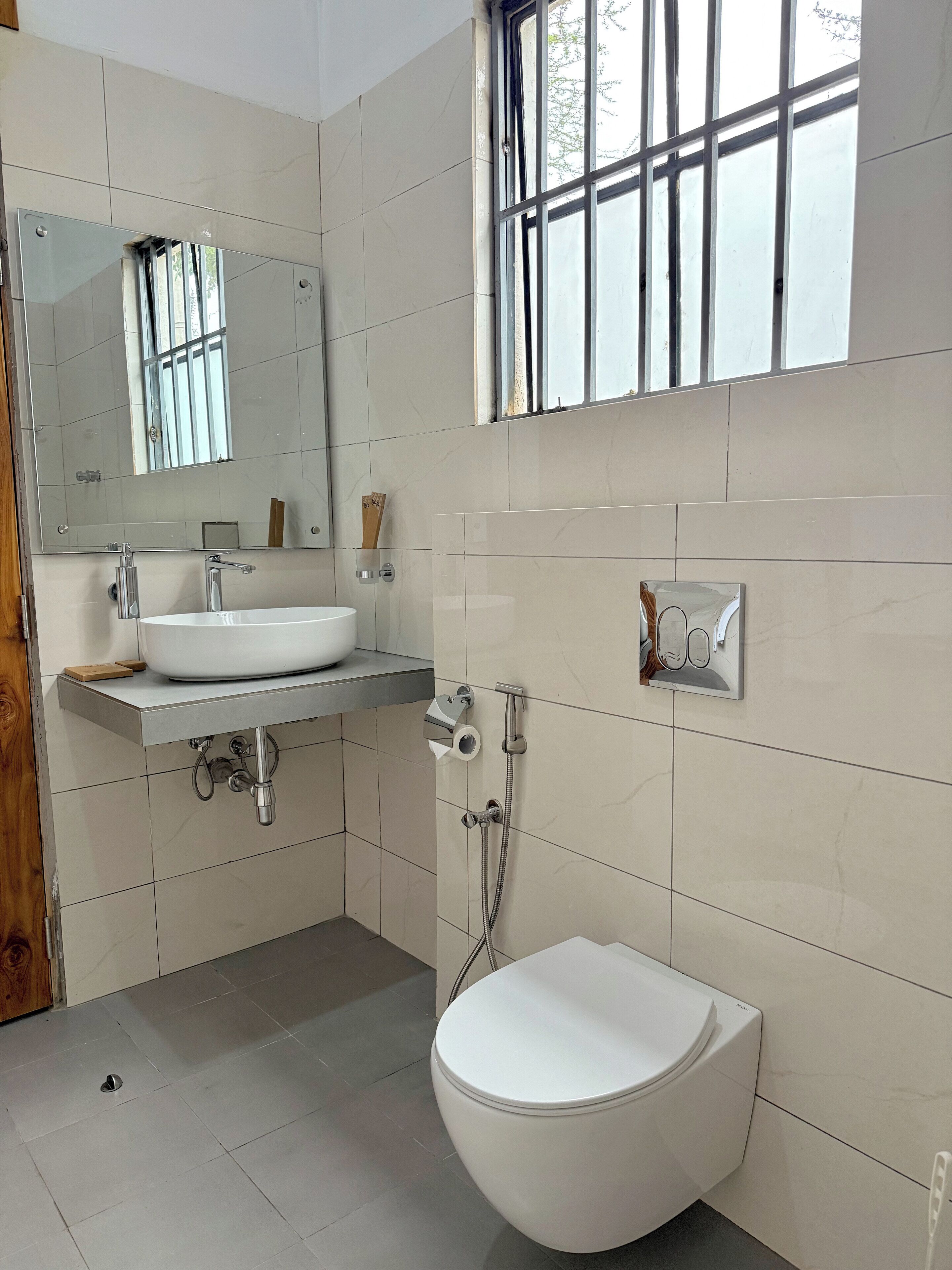 A modern bathroom with a white sink, mirror, and toilet. There is a window with bars, and the walls are tiled.