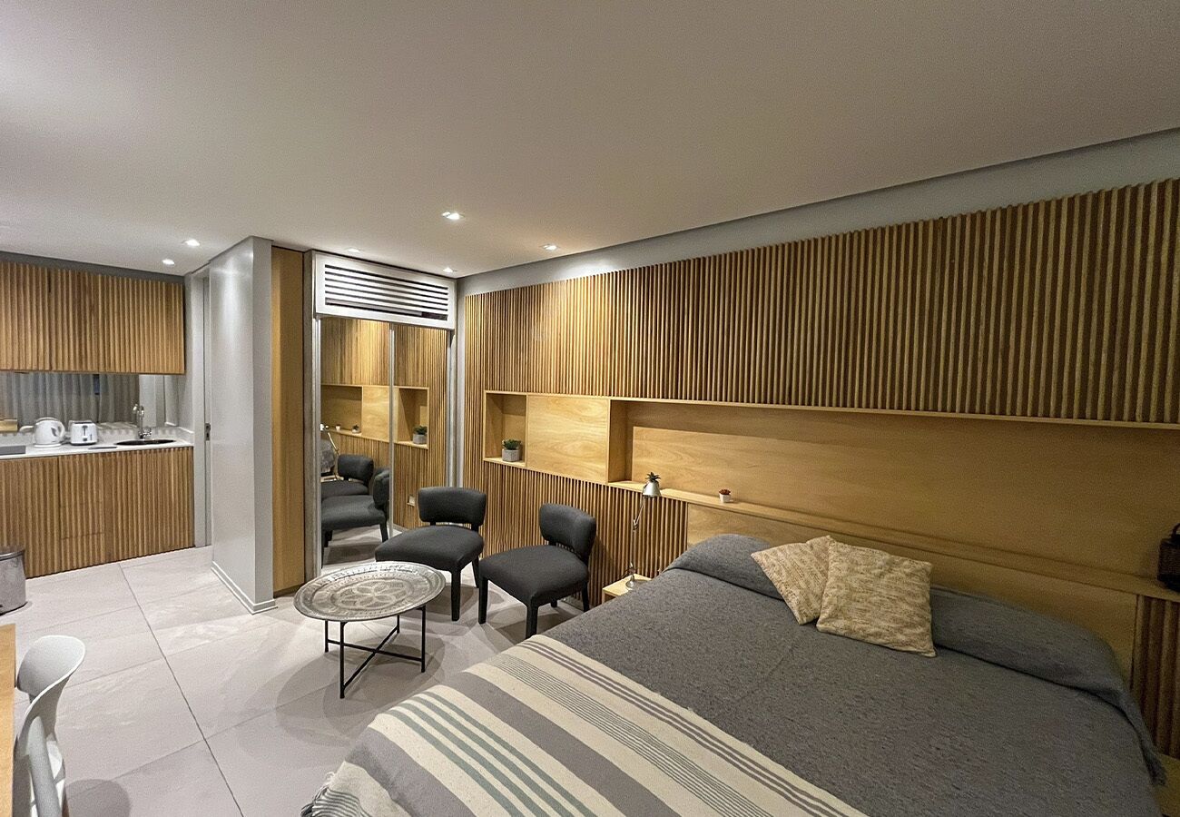 A modern hotel room with a large bed, a small round table, two armchairs, a kitchenette, and a wooden headboard.
