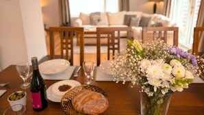 Uma mesa de jantar com uma garrafa de vinho, um pão, uma tigela de azeitonas e um vaso de flores.