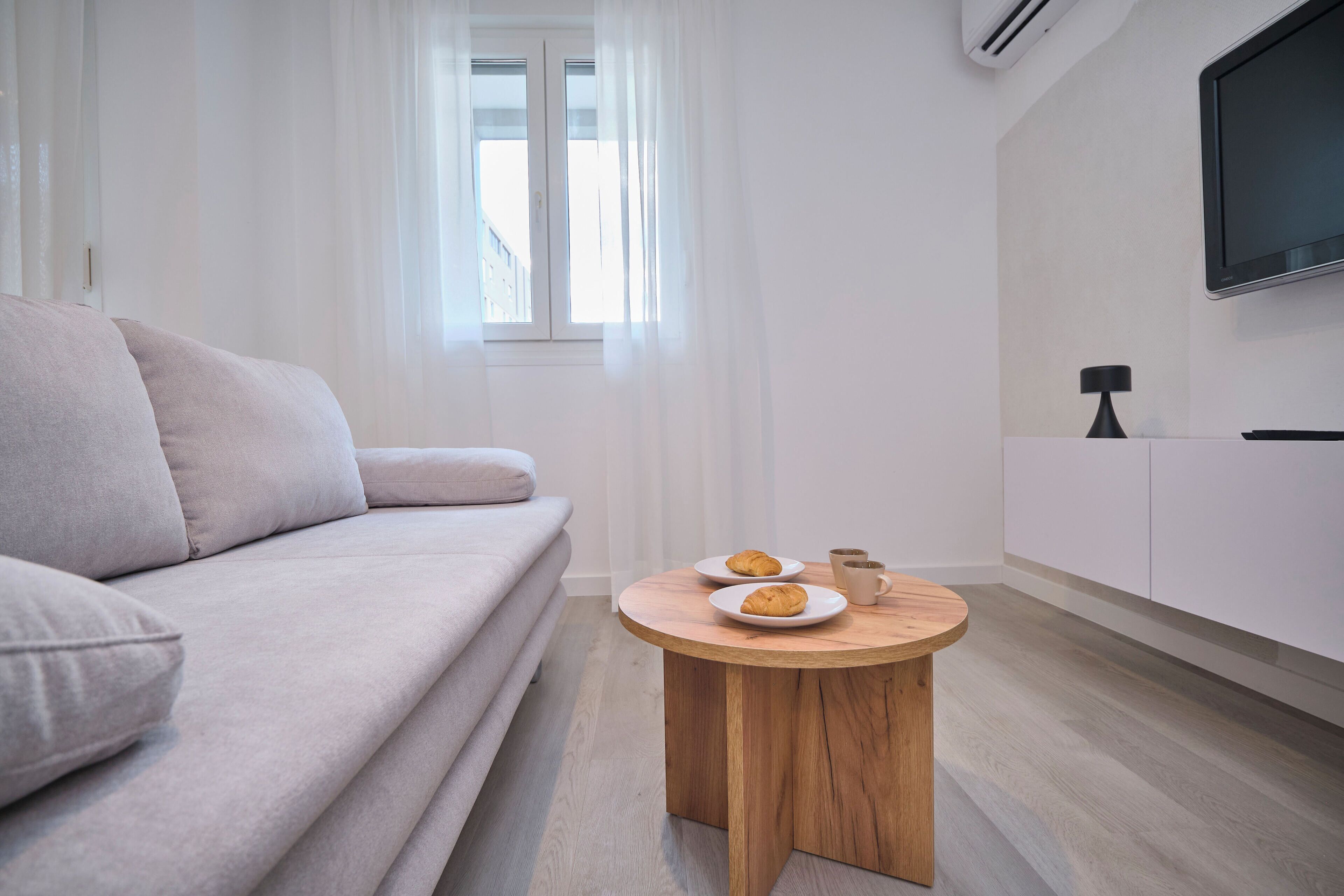 Een moderne woonkamer met een bank, een ronde houten salontafel met croissants, een kopje en een televisie aan de muur.