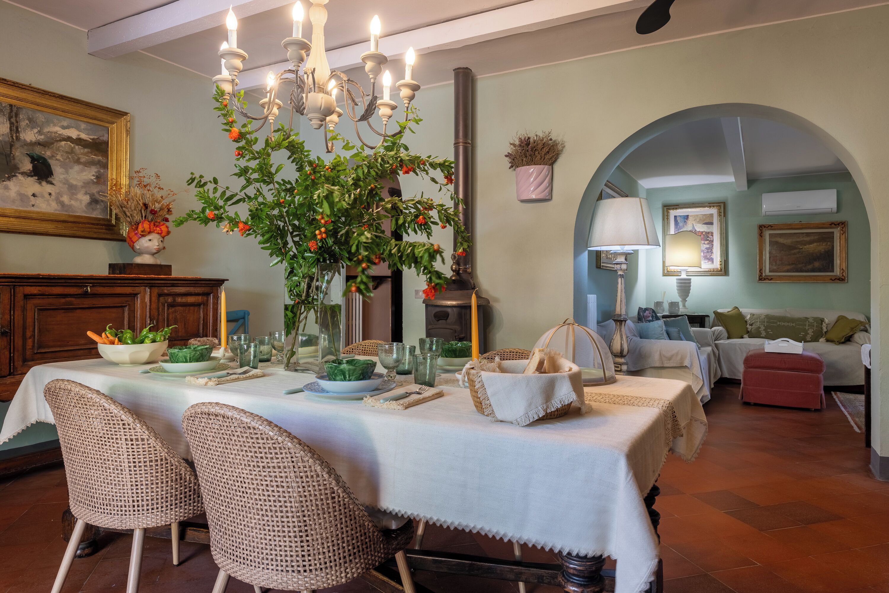 A dining area with a table set for four, surrounded by wicker chairs, a chandelier, and a vase with flowers.