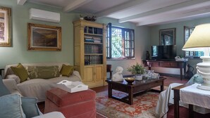 A living room with a sofa, ottoman, coffee table, bookshelf, and a television. - Podere la Botte Pool Lari (Lari)