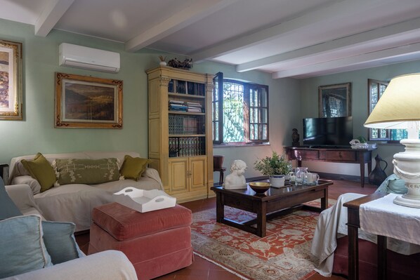 A living room with a sofa, ottoman, coffee table, bookshelf, and a television. - Podere la Botte Pool Lari (Lari)