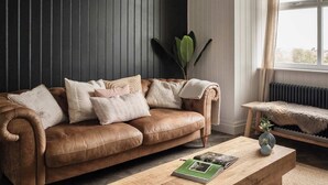 A brown leather sofa with multiple cushions, a wooden coffee table with a book and a vase, and a large window with curtains. - Whole Coastal Estate · Sleeps 12 · Anglesey (Isle of Anglesey)