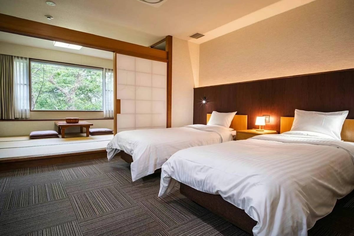 A hotel room with two beds, a small table, and a window with a view of greenery.