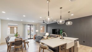 A modern kitchen with a central island, a dining area with a wooden table and chairs, and a living area with a sofa and TV. - Next to Pool, Sleek & Modern 3BR w/Fireplace (St. George)