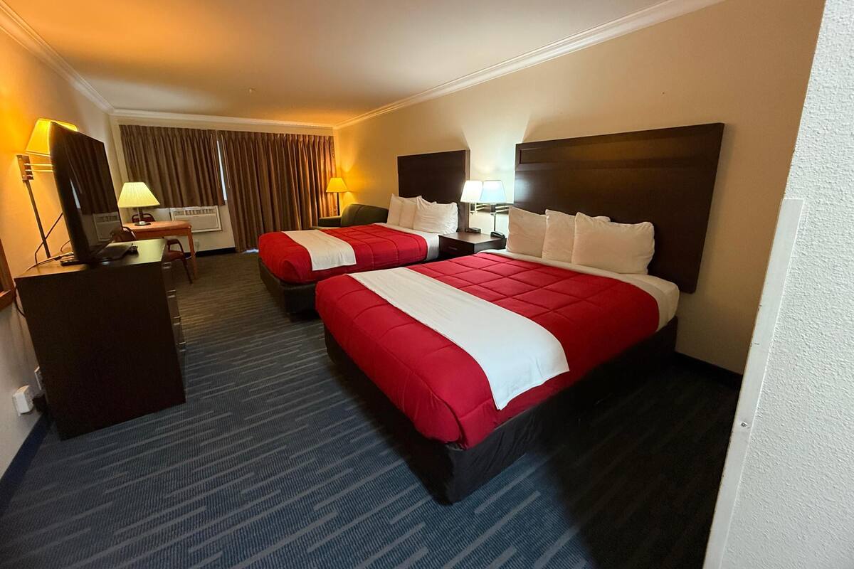 A hotel room with two beds, a desk, a chair, a lamp, and a television.