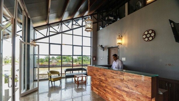 A reception area with a wooden counter, a seating area with chairs and a table, and a large window providing a view of greenery outside. - Hotel The Cult (Hubli)
