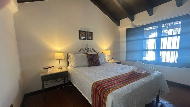 Un dormitorio con cama, dos mesitas de noche con lámparas, una ventana con persianas y un techo con vigas a la vista.