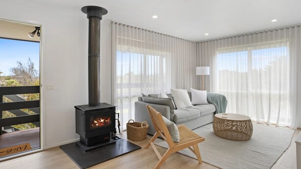 Uma sala de estar moderna com uma lareira a lenha, um sofá cinza, uma cadeira de madeira e uma grande janela com cortinas leves.
