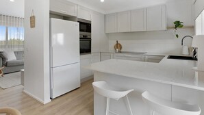 Uma cozinha moderna com armários brancos, uma ilha central com banquetas, forno embutido e geladeira.