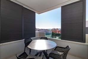 En balkon med et rundt bord og stole, store vinduer med skodder, og udsigt til bygninger i det fjerne.