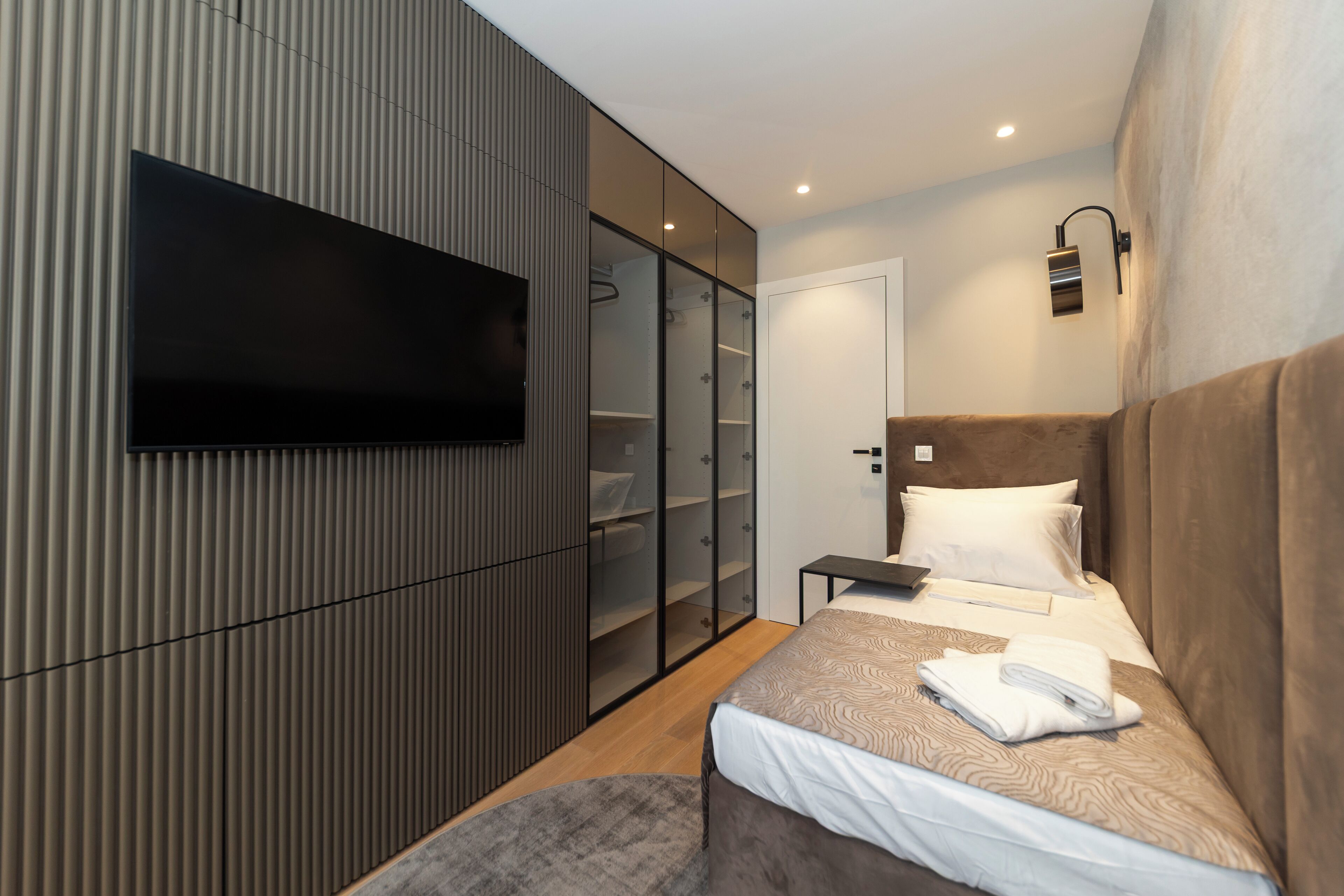 A modern hotel room with a large bed, a flat-screen TV mounted on the wall, a wardrobe with glass doors, and a bedside table.