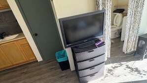 Una habitación con televisión sobre un mueble bajo, una puerta, un cubo de basura, un armario pequeño y una ventana con cortinas.