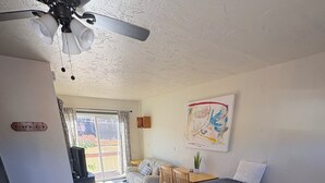 Un dormitorio con cama, ventilador de techo, ventana con cortinas, una mesita y una obra de arte colgada en la pared.