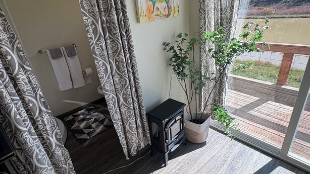 Una habitación con cortinas estampadas, un cuadro, una planta en maceta, una estufa negra pequeña y vistas a un bosque a través de un ventanal.