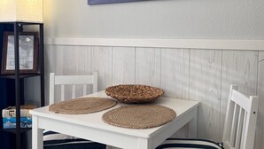 Una habitación con una mesa blanca, dos sillas, un mantel tejido, un certificado enmarco y un cuadro colgado en la pared con una escena de playa.