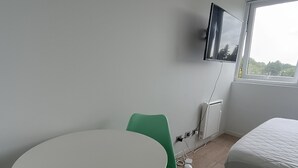 Une pièce avec une table ronde blanche, deux chaises vertes, un lit recouvert d’une couette blanche, un téléviseur fixé au mur et une fenêtre donnant sur des arbres.