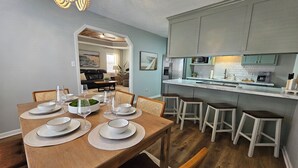 Ein Essbereich mit Holztisch, gedeckt für vier Personen, eine Küche mit Marmorarbeitsplatte und ein Wohnzimmer im Hintergrund.