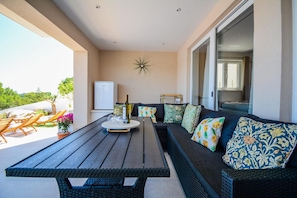 A sunlit patio with a wooden table, a sofa with patterned cushions, and a decorative wall piece.