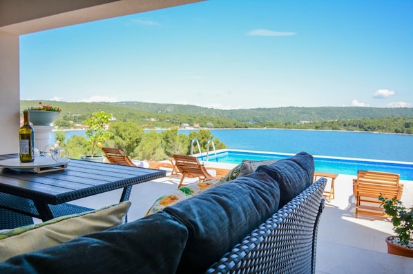 A balcony with a view of a large body of water, a pool, and a dining area with outdoor furniture.