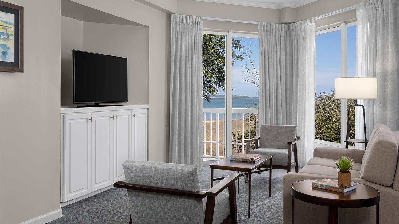 Ein Hotelzimmer mit Flachbildfernseher, einer Couch, einem Sessel, einem kleinen Tisch und Meerblick durch große Fenster.