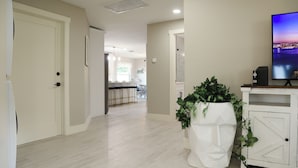 A hallway with a white door, a potted plant, a TV on a white cabinet, and a kitchen in the background. - Modern Coastal Villa • Private Fenced Yard • Minutes to the Beach (Naples)