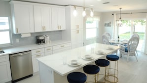 A modern kitchen with white cabinetry, a central island with stools, and a dining area with wicker chairs. - Modern Coastal Villa • Private Fenced Yard • Minutes to the Beach (Naples)