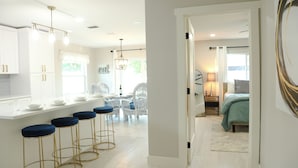 A modern kitchen with white cabinets, a bar area with blue stools, and a dining area with wicker chairs. - Modern Coastal Villa • Private Fenced Yard • Minutes to the Beach (Naples)