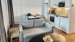 Ett modernt vardagsrum med en grå soffa, ett litet matbord och en köksdel med vita skåp och vitvaror.
