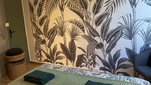 Une chambre avec un lit, un ventilateur de plafond, un mur orné d’une fresque représentant des feuilles tropicales, une porte verte et un panier en osier.